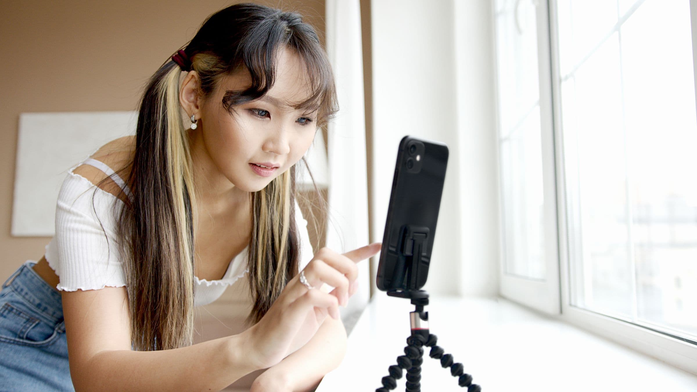 A woman setting up a phone on a small tripod beside a bright window, ready to film.